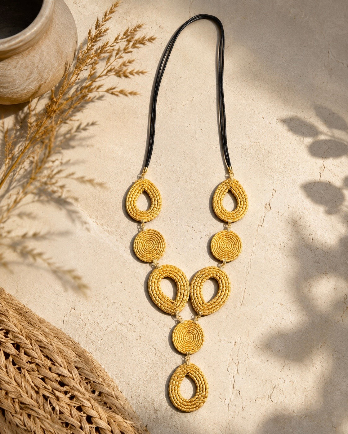 Collana maxi in Capim Dourado con elementi geometrici intrecciati a mano e cordoncino nero, ambientata su sfondo naturale dai toni sabbia, realizzata in autentico oro vegetale brasiliano.