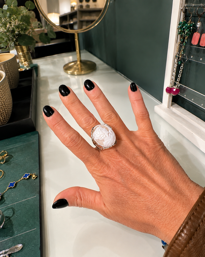 Anello cameo in conchiglia naturale inciso con profilo femminile, indossato su mano con manicure nera in elegante ambiente boutique gioielleria.