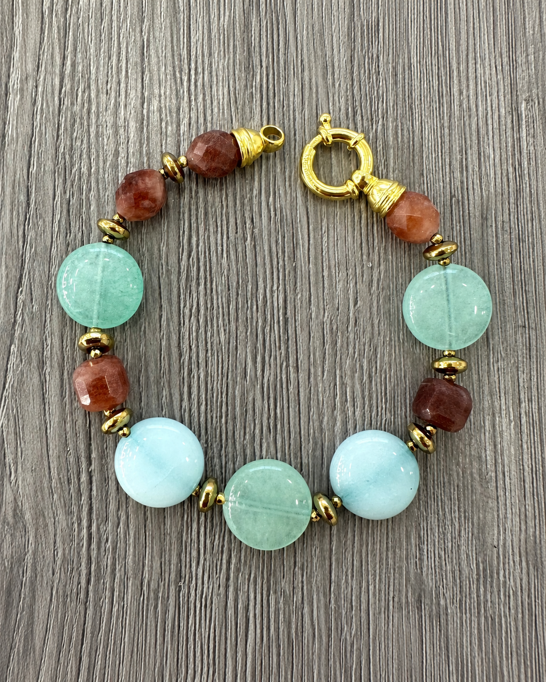 Bracciale aperto in argento dorato con Tormalina, Giada Verde e Azzurra ed Ematite dorata, con chiusura tonda visibile su legno grigio.