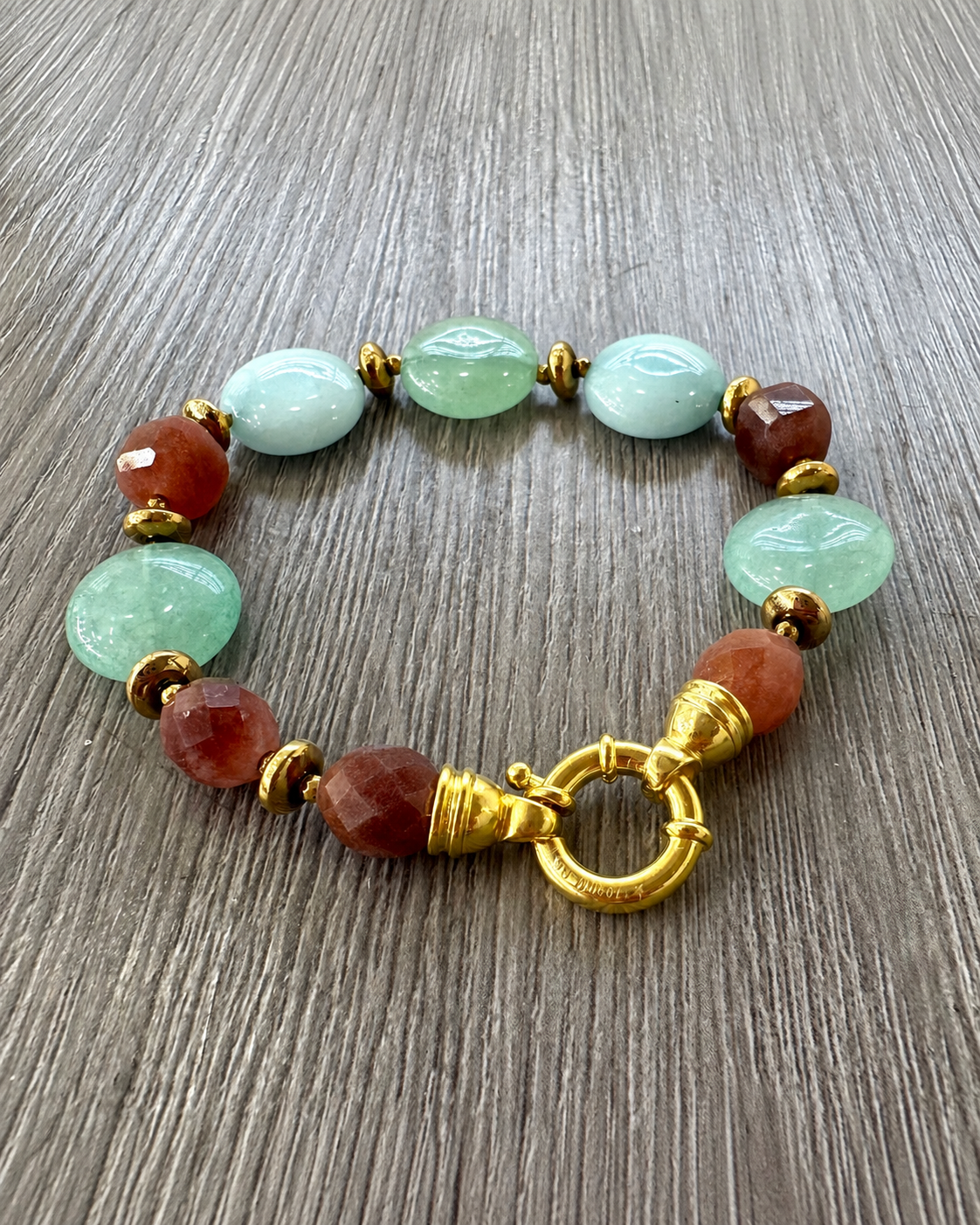 Bracciale in argento dorato con giada verde e azzurra, tormalina sfaccettata ed ematite dorata, con chiusura tonda, su legno grigio.
