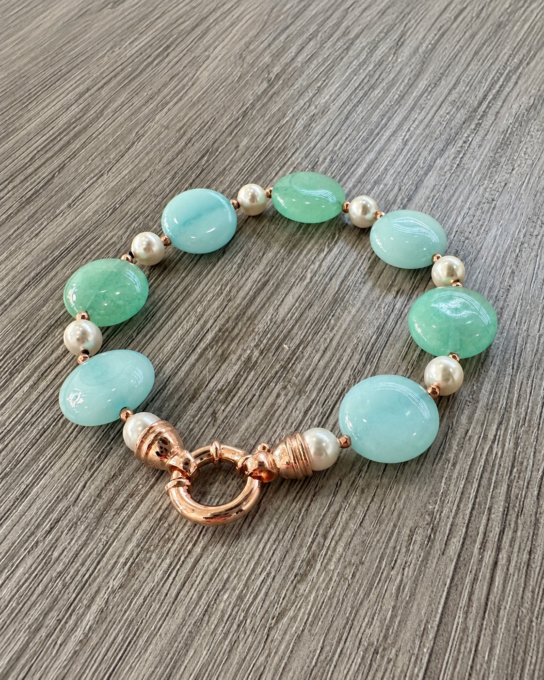 Bracciale in argento rosato con perle Maiorca, giada verde e azzurra e chiusura tonda, appoggiato su legno grigio.