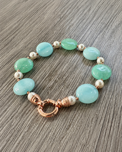 Bracciale in argento rosato con perle Maiorca, giada verde e azzurra e chiusura tonda, appoggiato su legno grigio.