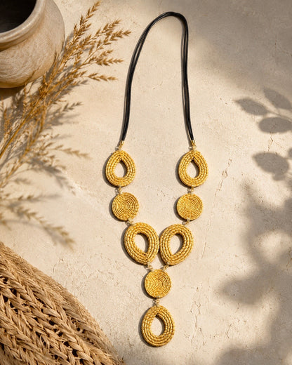 Collana maxi in Capim Dourado con elementi geometrici intrecciati a mano e cordoncino nero, ambientata su sfondo naturale dai toni sabbia, realizzata in autentico oro vegetale brasiliano.