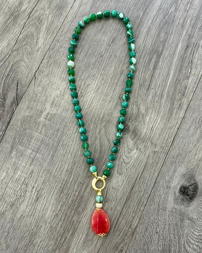     Collana artigianale in Agata Brasiliana striata verde con pendente in Giada rosa, dettagli in Argento 925 dorato con zirconi ed Ematite dorata, fotografata su fondo effetto legno.