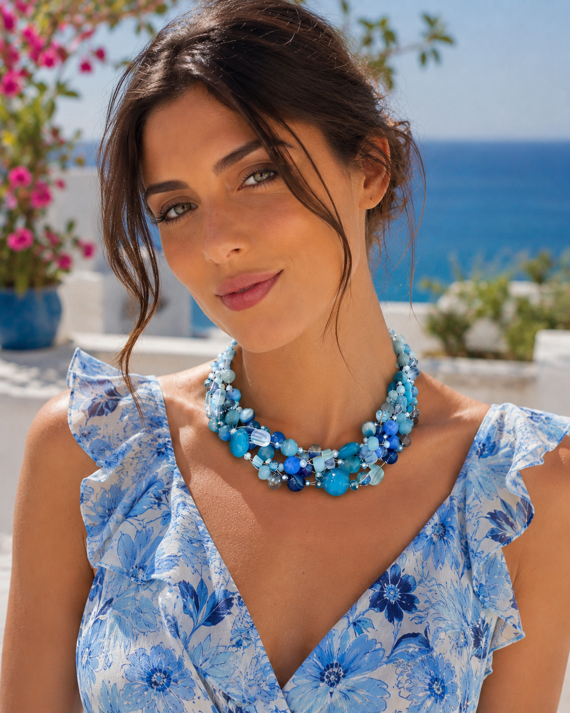 Collana artigianale multifilo blu e azzurra indossata da modella con abito floreale estivo nelle tonalità del celeste e del blu, ambientazione elegante sul mare.