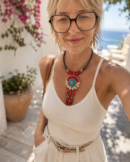 Collana boho etnica con perline rosse, turchesi e dettagli pendenti indossata da una giovane modella con outfit estivo chiaro in ambientazione mediterranea luminosa.