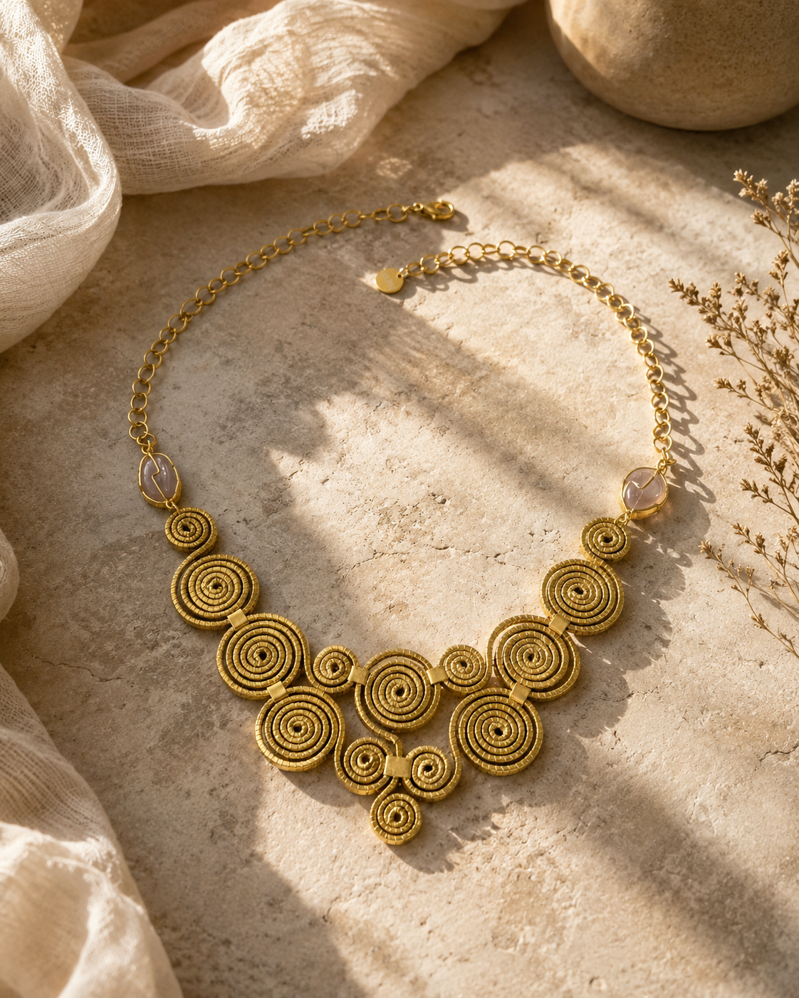 Collana Capim Dourado con spirali dorate e pietra viola fotografata in ambientazione naturale beige con luce calda.