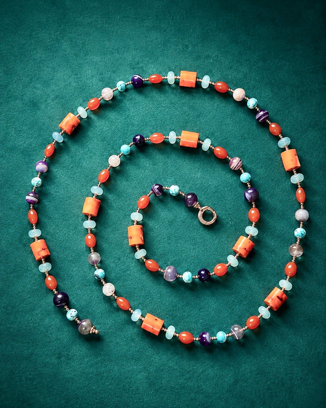 Collana lunga artigianale con corallo bambù, giada azzurra, agata viola e dettagli in argento rosato, fotografata su fondo verde petrolio.