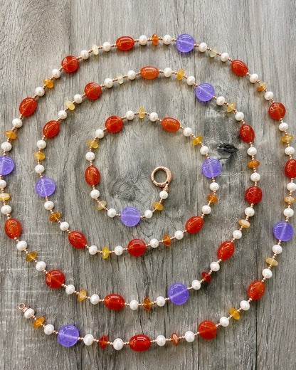     Collana artigianale lunga con Giada viola, Giada arancio, Corniola, Perle ed Ematite rosata fotografata su fondo effetto legno.