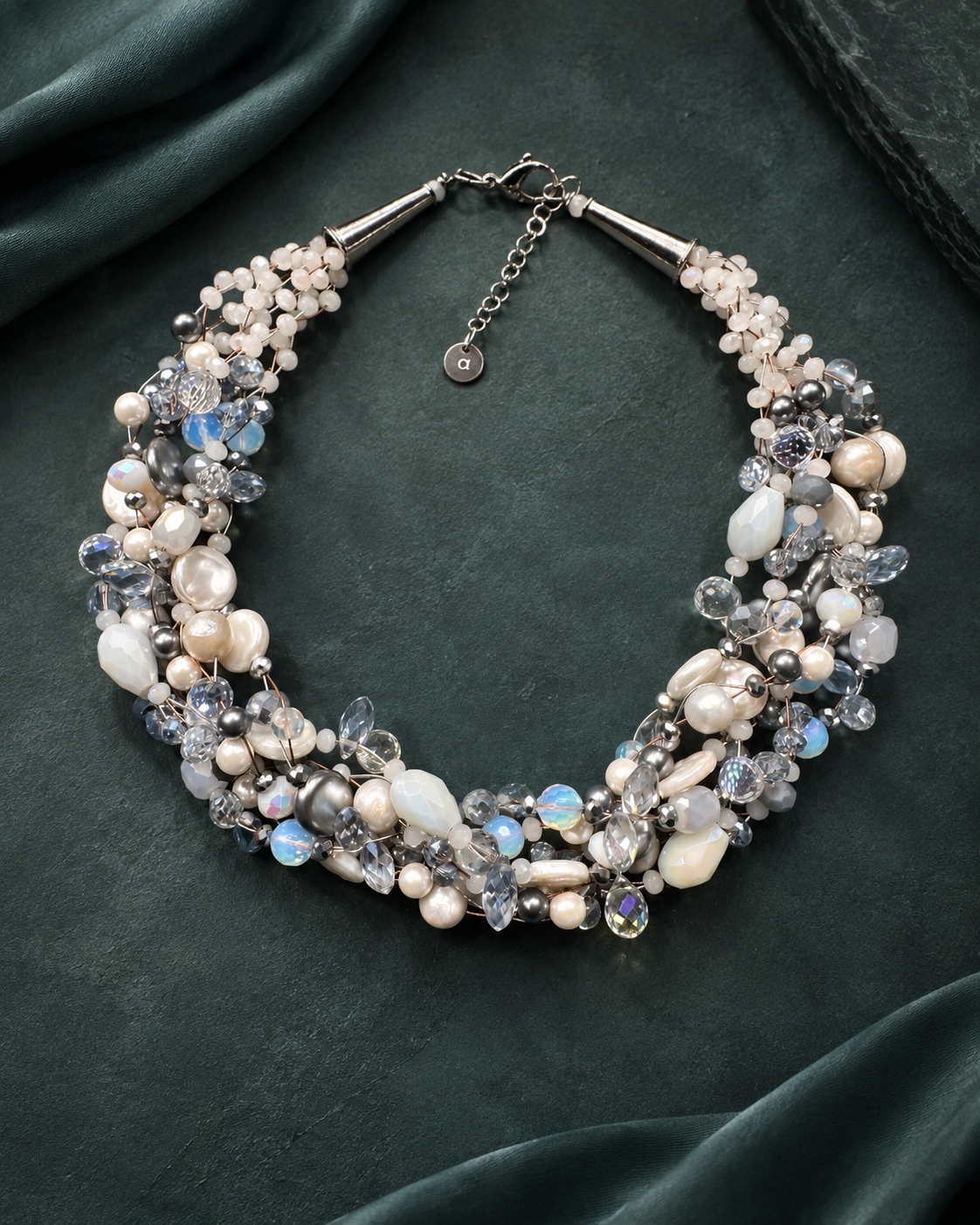 Collana intrecciata color ghiaccio con perle e cristalli nelle sfumature del bianco, grigio e azzurro, finitura argentata, fotografata su fondo verde petrolio scuro.