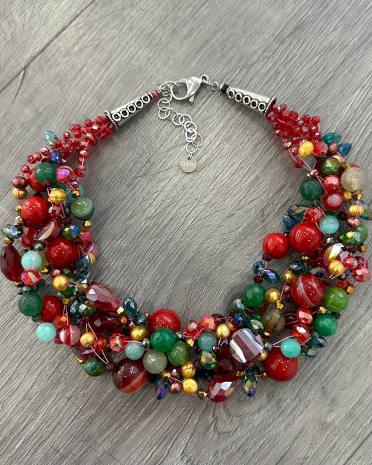 Collana intrecciata multicolor con perle rosse, verdi e cristalli fotografata su sfondo legno naturale.