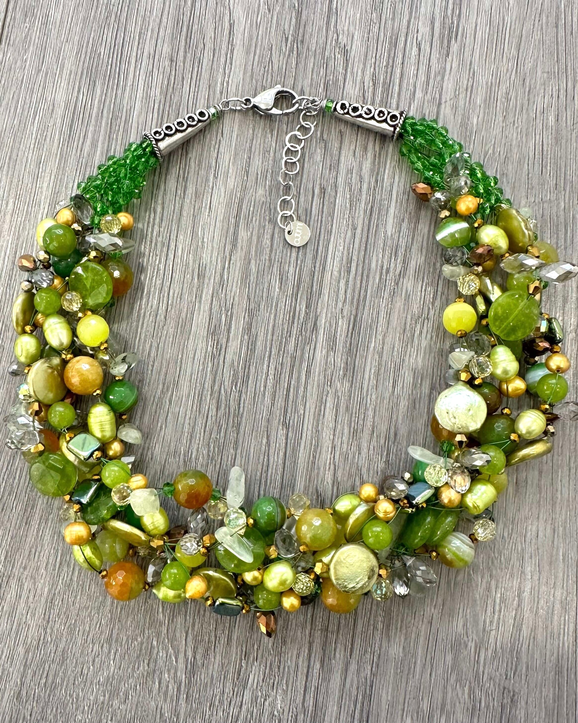Collana intrecciata artigianale con pietre verdi lime,perle e cristalli appoggiata su superficie legno grigio￼