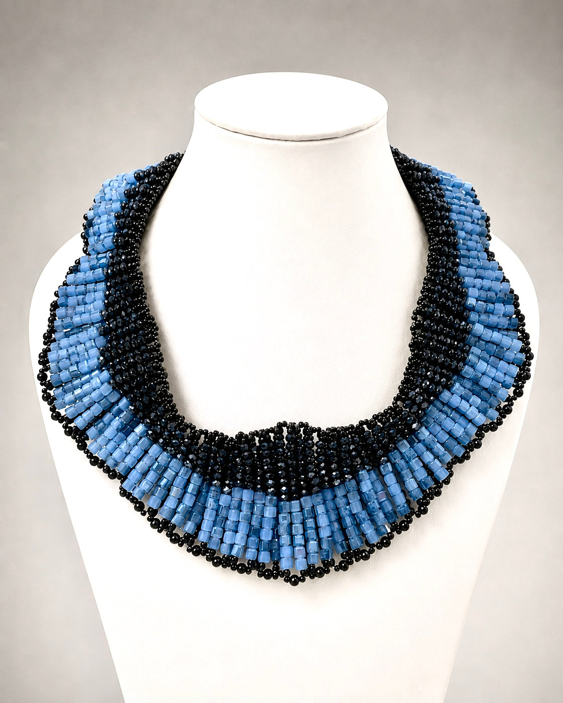 Collana ricamata a mano con perline blu e nere e cristalli esposta su busto bianco.