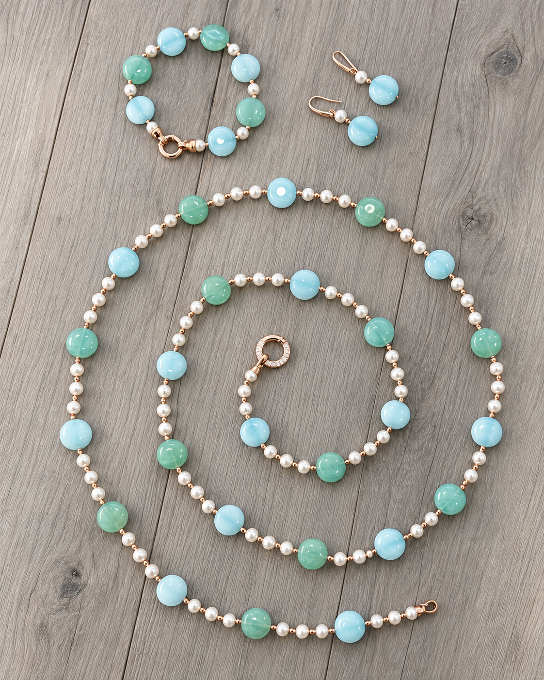 Parure in argento rosato con perle e giada verde e azzurra: collana con chiusura tonda, bracciale e orecchini su fondo in legno grigio.
